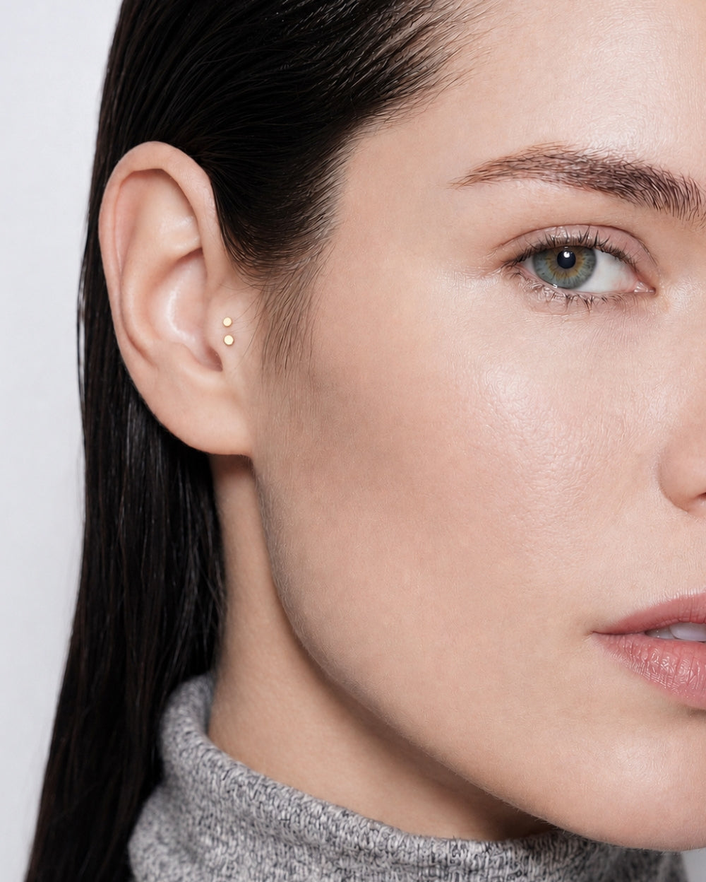 Close-up of a woman's ear wearing a delicate gold micro-piercing stud earring, with her face and dark hair visible against a light gray background.