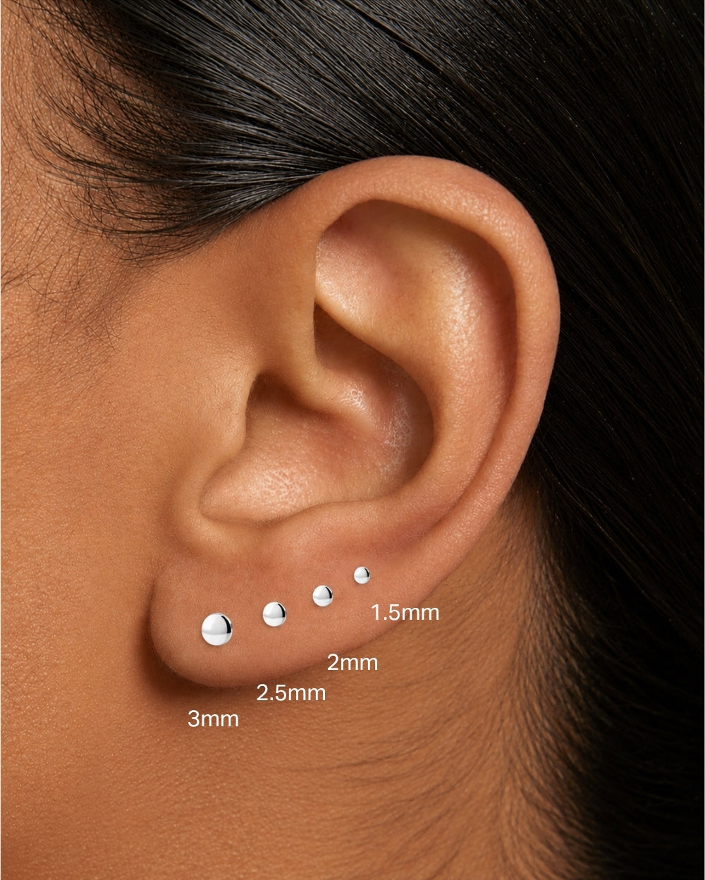 Close-up of a person's ear showing four small silver ear studs placed on the lobe, with size labels reading 3mm, 2.5mm, 1.