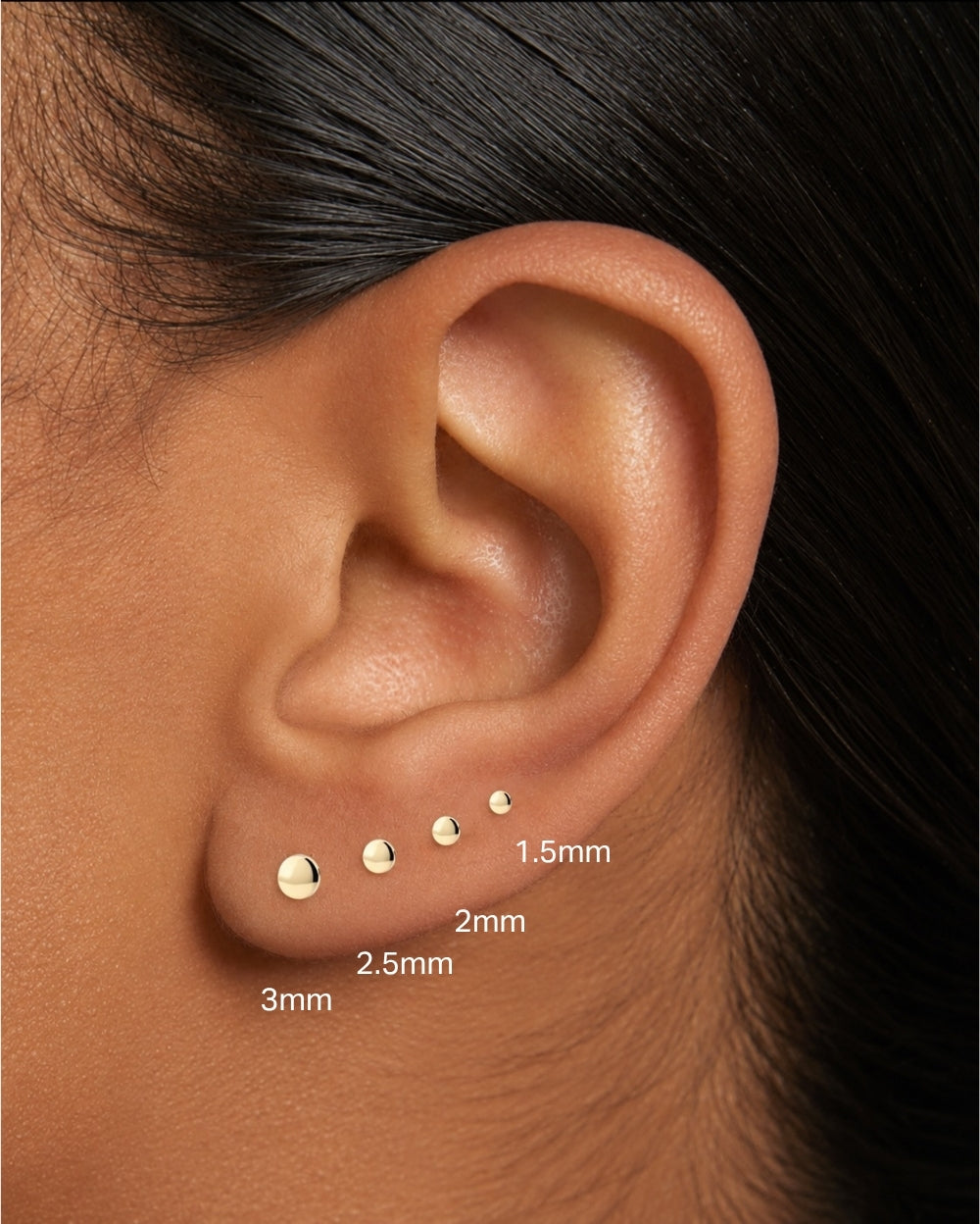 Close-up of a person's ear showing three small gold ear piercings at different sizes labeled 3mm, 2.5mm, 2mm, and 1.