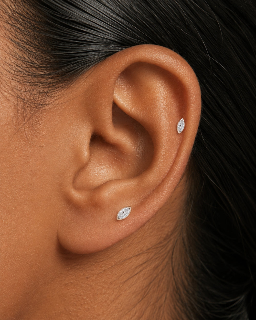 Close-up of a person's ear wearing two small white gold marquise diamond stud earrings, one at the lobe and one at the helix, with dark hair visible.