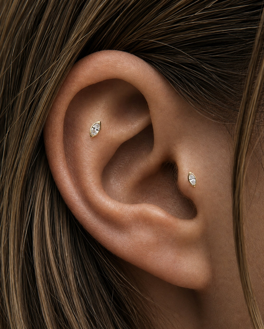 Close-up of a person's ear wearing two small marquise-cut stud earrings in gold settings, with brown hair framing the ear.
